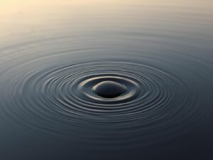 A serene natural scene with still water reflecting the sky, a single stone creating gentle ripples that slowly expand outward